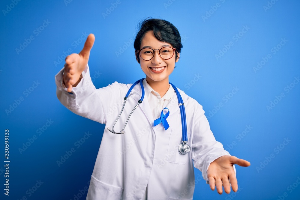 Young beautiful asian doctor girl wearing stethoscope and coat with blue cancer ribbon looking at the camera smiling with open arms for hug. Cheerful expression embracing happiness.
