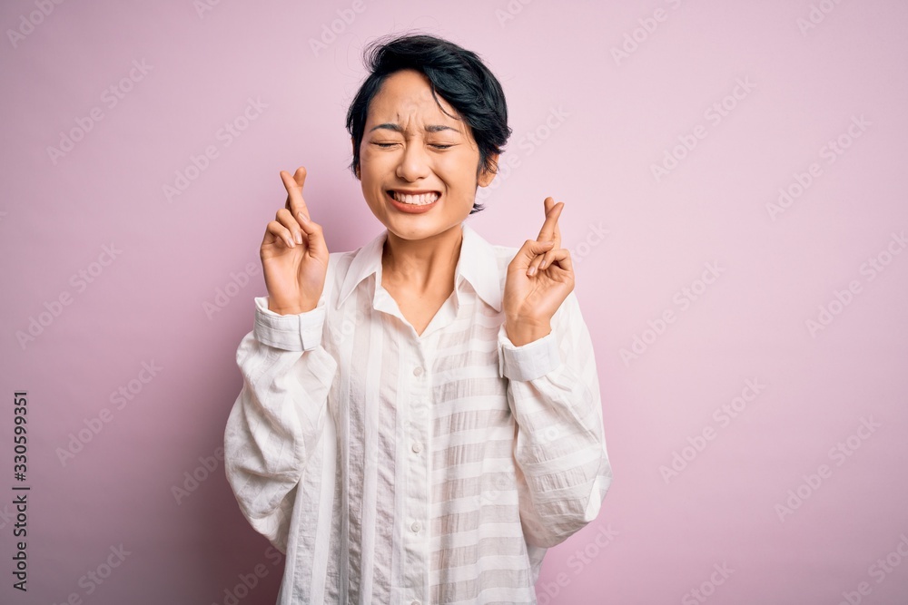 Young beautiful asian girl wearing casual shirt standing over isolated pink background gesturing finger crossed smiling with hope and eyes closed. Luck and superstitious concept.