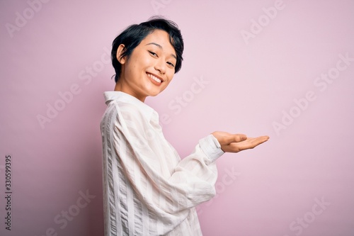 Young beautiful asian girl wearing casual shirt standing over isolated pink background pointing aside with hands open palms showing copy space, presenting advertisement smiling excited happy