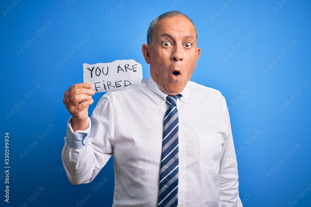 Senior handsome businessman holding you are fired paper over isolated blue background scared in shock with a surprise face, afraid and excited with fear expression Senior handsome businessman holding you are fired paper over isolated blue background scared in shock with a surprise face, afraid and excited with fear expression