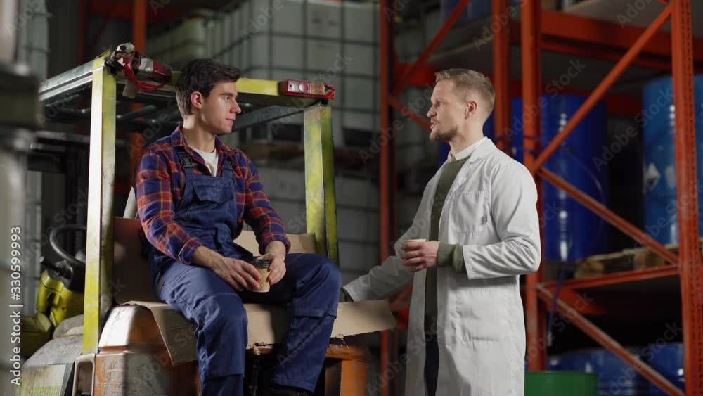 Tracking medium shot of middle aged warehouse worker in white coat talking to young forklift driver during coffee break in industrial storage
