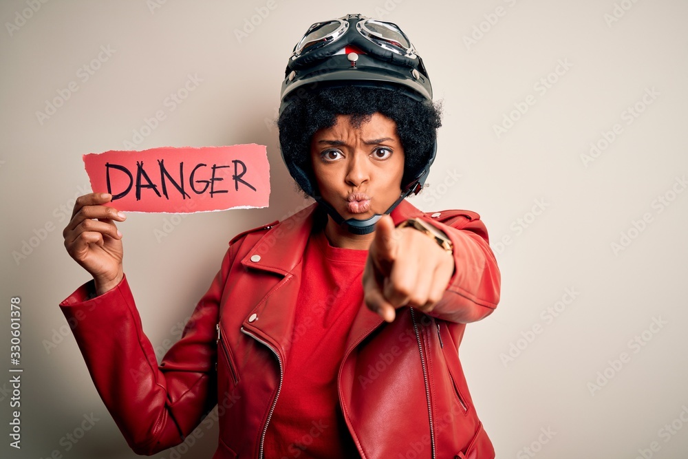 Young African American afro motorcyclist woman with curly hair holding danger paper message pointing with finger to the camera and to you, hand sign, positive and confident gesture from the front Young African American afro motorcyclist woman with curly hair holding danger paper message pointing with finger to the camera and to you, hand sign, positive and confident gesture from the front