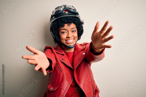 Young African American afro motorcyclist woman with curly hair wearing motorcycle helmet looking at the camera smiling with open arms for hug. Cheerful expression embracing happiness.