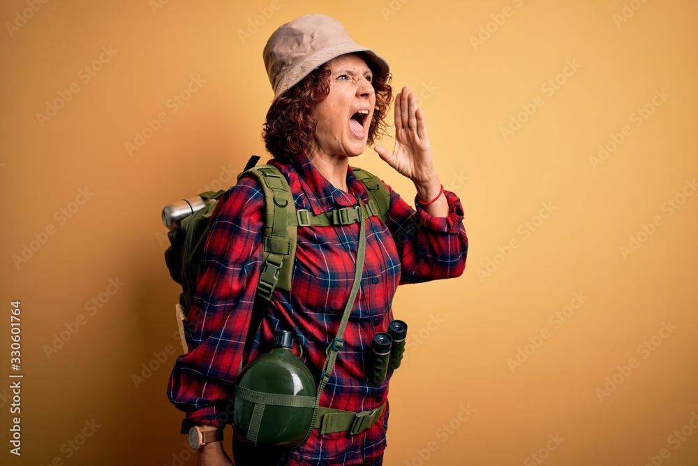Middle age curly hair hiker woman hiking wearing backpack and water canteen using binoculars shouting and screaming loud to side with hand on mouth. Communication concept.