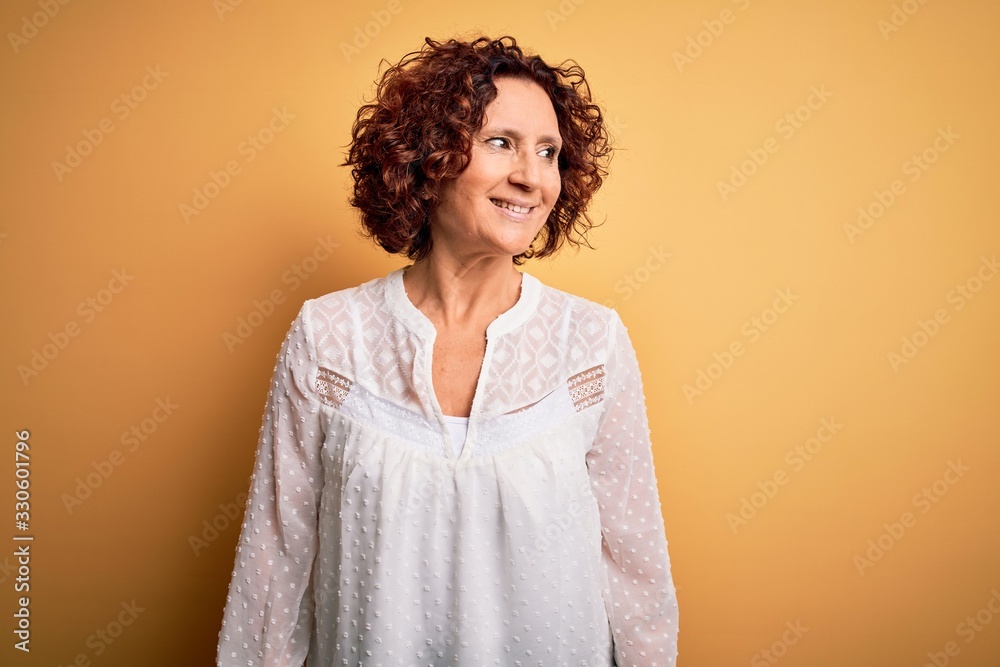 Middle age beautiful curly hair woman wearing casual summer dress over yellow background looking away to side with smile on face, natural expression. Laughing confident. Middle age beautiful curly hair woman wearing casual summer dress over yellow background looking away to side with smile on face, natural expression. Laughing confident.