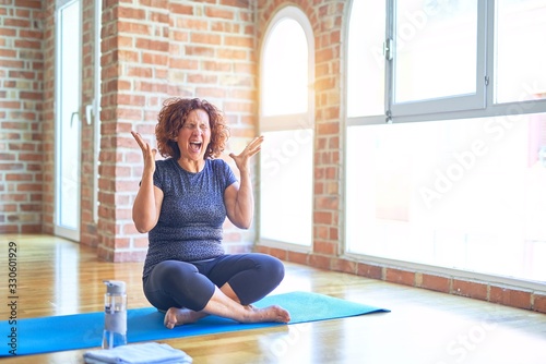 Middle age beautiful sportswoman wearing sportswear sitting on mat practicing yoga at home celebrating mad and crazy for success with arms raised and closed eyes screaming excited. Winner concept