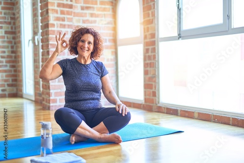Middle age beautiful sportswoman wearing sportswear sitting on mat practicing yoga at home smiling positive doing ok sign with hand and fingers. Successful expression.