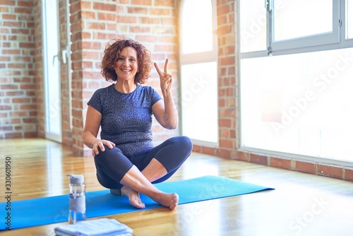 Middle age beautiful sportswoman wearing sportswear sitting on mat practicing yoga at home showing and pointing up with fingers number two while smiling confident and happy.