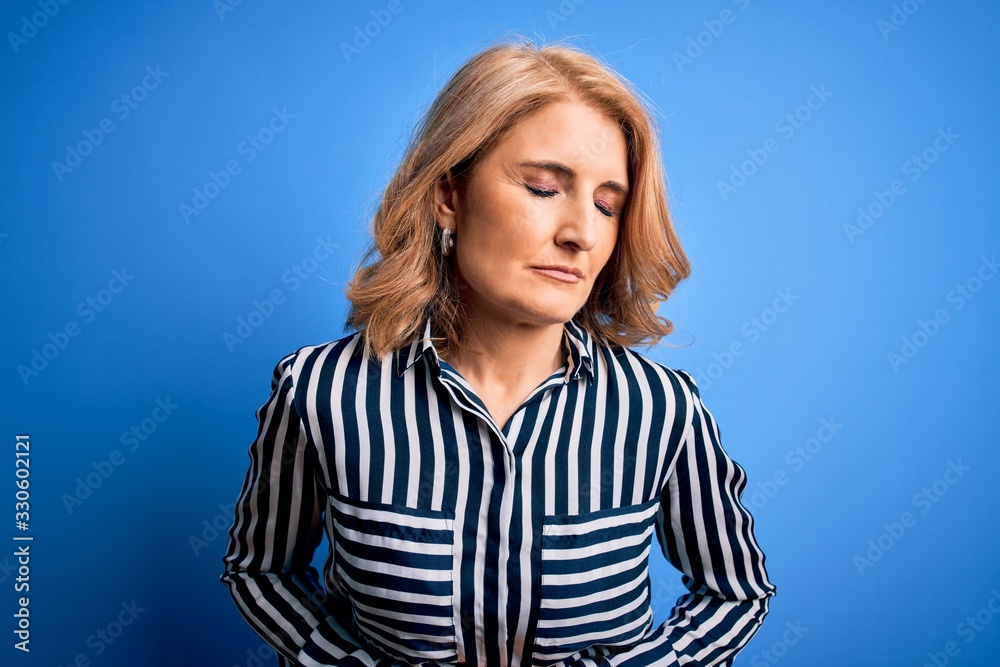 Middle age beautiful blonde woman wearing casual striped shirt standing over blue background with hand on stomach because indigestion, painful illness feeling unwell. Ache concept.