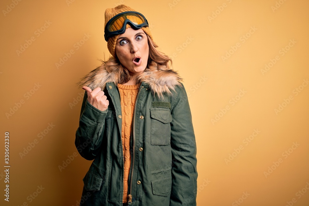 Middle age beautiful blonde skier woman wearing snow sportwear and ski goggles Surprised pointing with hand finger to the side, open mouth amazed expression.