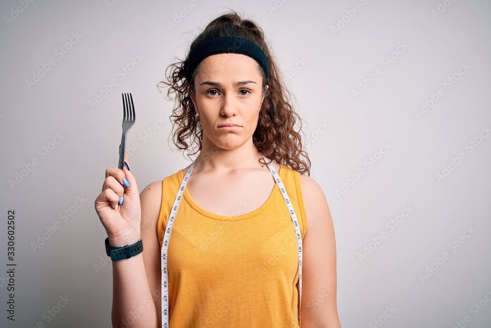 Young beautiful woman with curly hair doing diet holding fork and tape measure with a confident expression on smart face thinking serious Young beautiful woman with curly hair doing diet holding fork and tape measure with a confident expression on smart face thinking serious