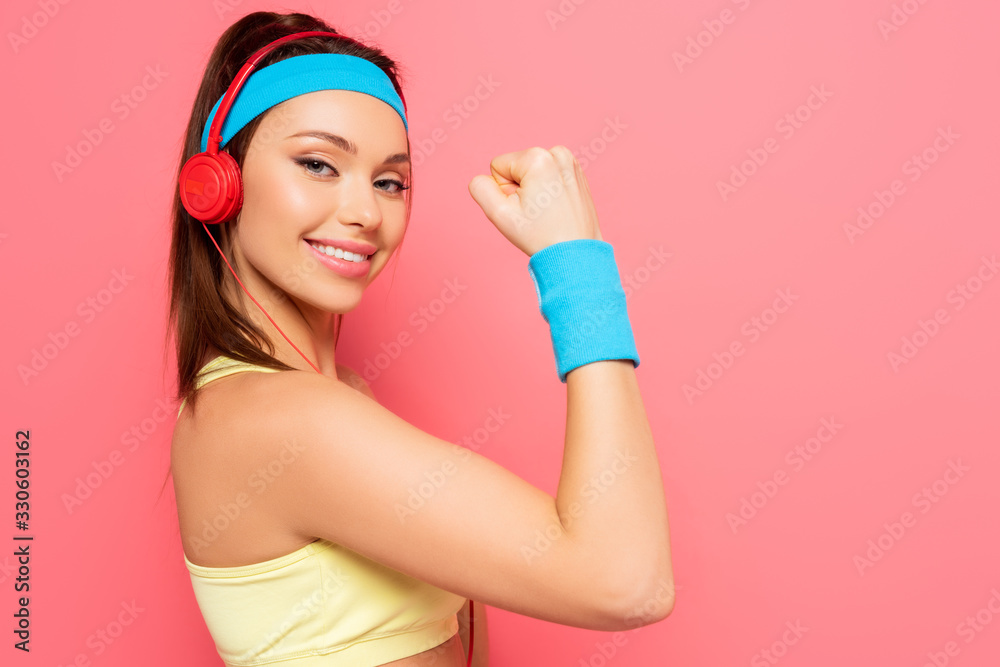 cheerful sportswoman in headphones demonstrating biceps while looking at camera on pink background cheerful sportswoman in headphones demonstrating biceps while looking at camera on pink background