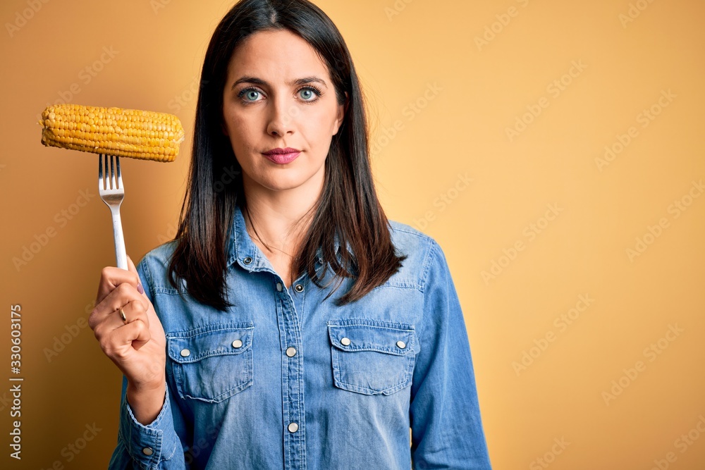 Young woman with blue eyes holding fork with fresh cob corn standing over yellow background with a confident expression on smart face thinking serious Young woman with blue eyes holding fork with fresh cob corn standing over yellow background with a confident expression on smart face thinking serious