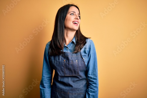 Young baker woman with blue eyes wearing apron standing over isolated yellow background looking away to side with smile on face, natural expression. Laughing confident. © Krakenimages.com