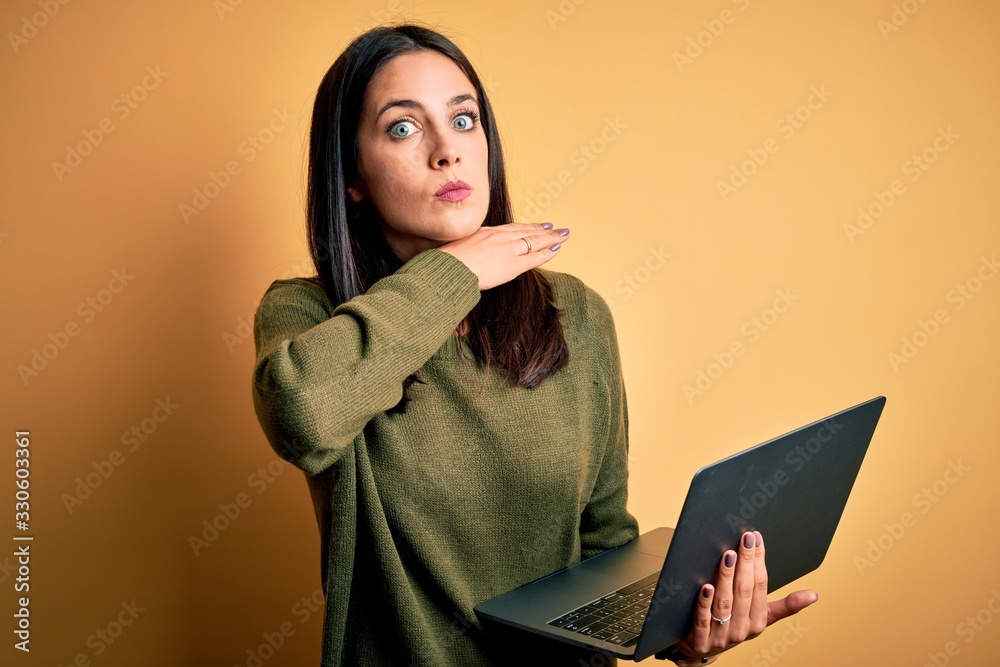 Young brunette woman with blue eyes working using computer laptop over yellow background cutting throat with hand as knife, threaten aggression with furious violence
