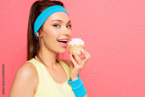 smiling sportswoman going to eat delicious cupcake while looking at camera isolated on pink