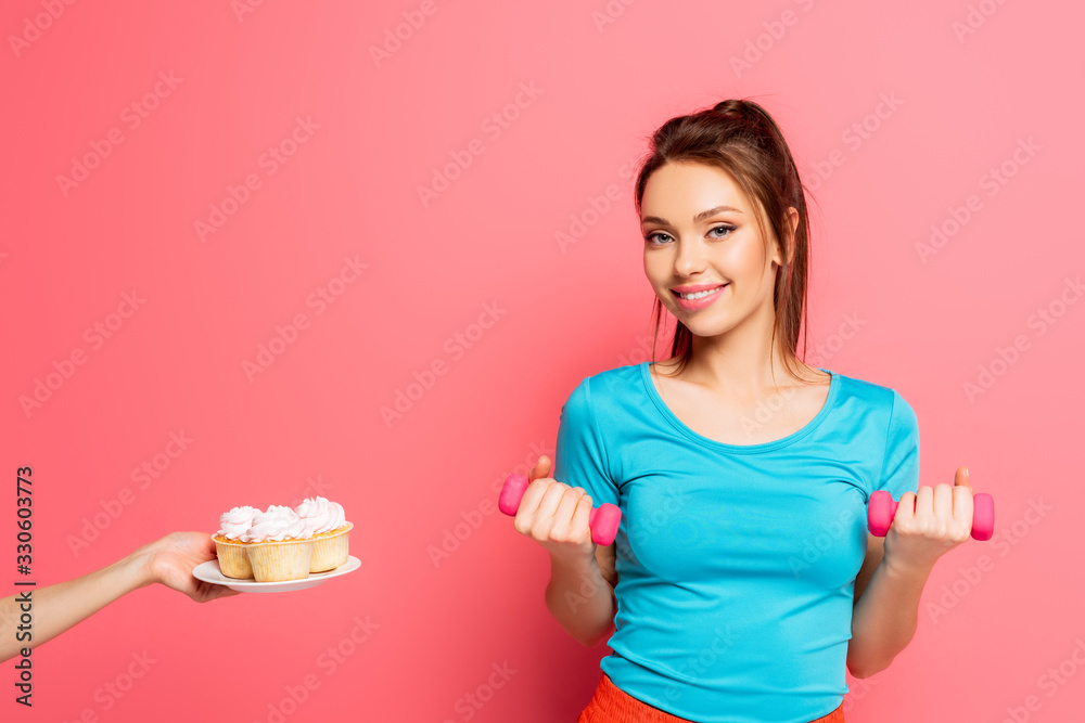 smiling sportswoman exercising with dumbbells near female hand with plate of delicious cupcakes on pink background smiling sportswoman exercising with dumbbells near female hand with plate of delicious cupcakes on pink background