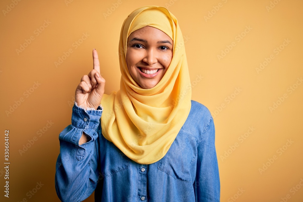 Young beautiful woman with curly hair wearing arab traditional hijab over yellow background showing and pointing up with finger number one while smiling confident and happy. Young beautiful woman with curly hair wearing arab traditional hijab over yellow background showing and pointing up with finger number one while smiling confident and happy.