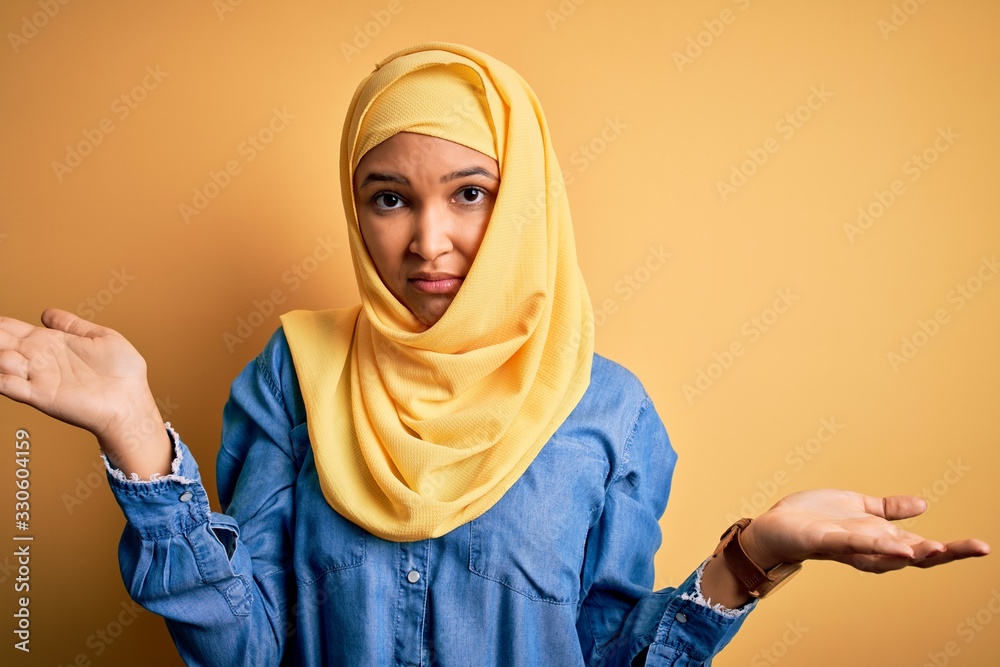 Young beautiful woman with curly hair wearing arab traditional hijab over yellow background clueless and confused with open arms, no idea concept. Young beautiful woman with curly hair wearing arab traditional hijab over yellow background clueless and confused with open arms, no idea concept.