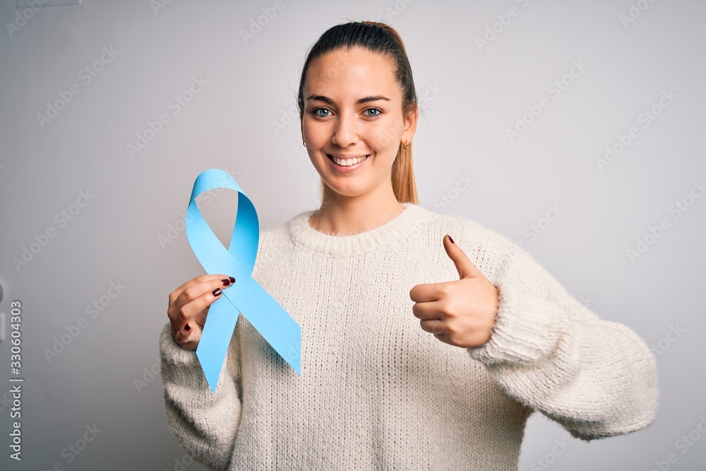 Young beautiful blonde woman holding blue cancer ribbon over isolated white background happy with big smile doing ok sign, thumb up with fingers, excellent sign Young beautiful blonde woman holding blue cancer ribbon over isolated white background happy with big smile doing ok sign, thumb up with fingers, excellent sign