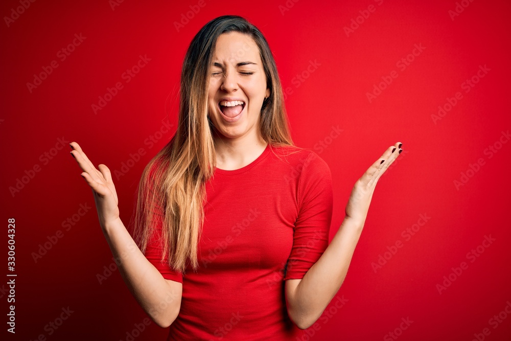 Young beautiful blonde woman with blue eyes wearing casual t-shirt over red background celebrating mad and crazy for success with arms raised and closed eyes screaming excited. Winner concept
