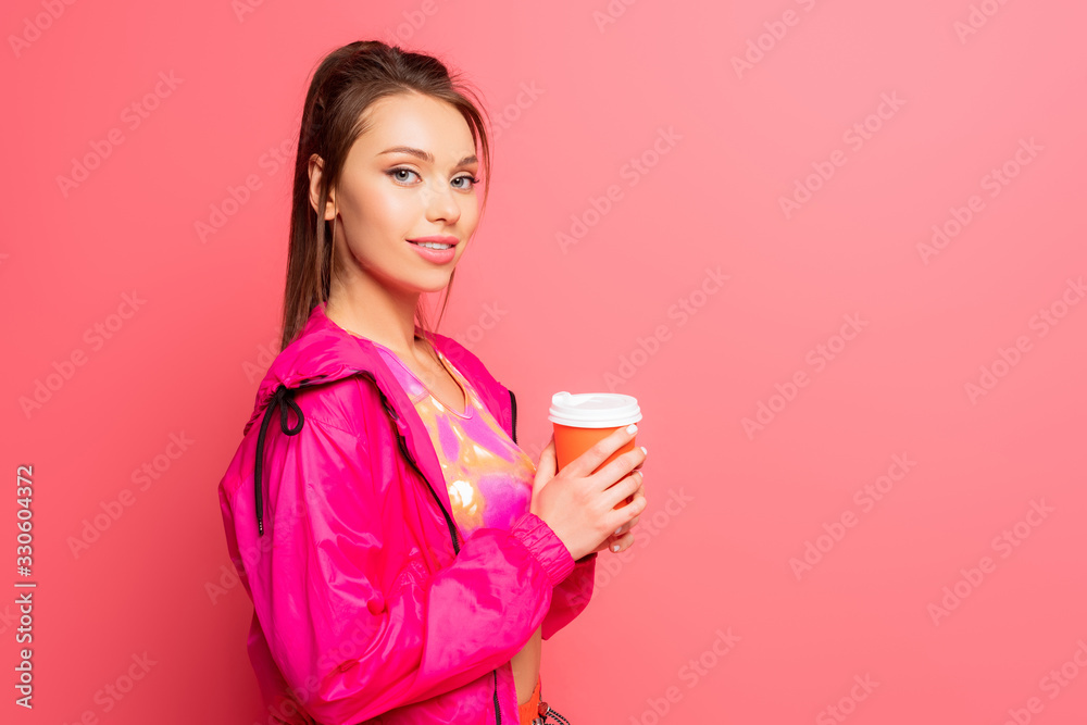 pretty, smiling sportswoman holding coffee to go while looking at camera on pink background pretty, smiling sportswoman holding coffee to go while looking at camera on pink background