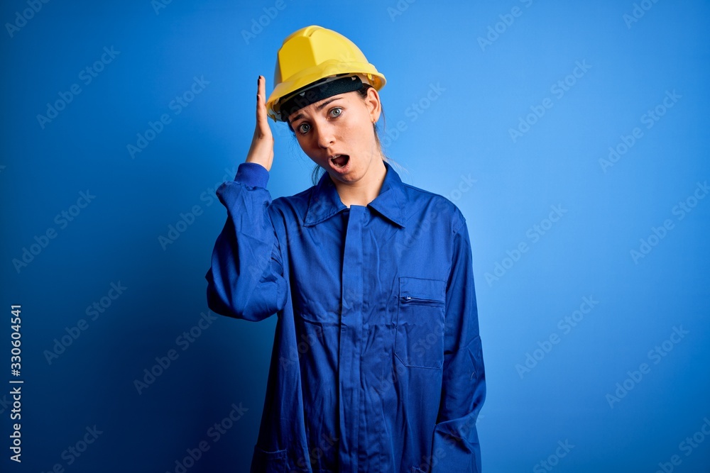 Young beautiful worker woman with blue eyes wearing security helmet and uniform surprised with hand on head for mistake, remember error. Forgot, bad memory concept.