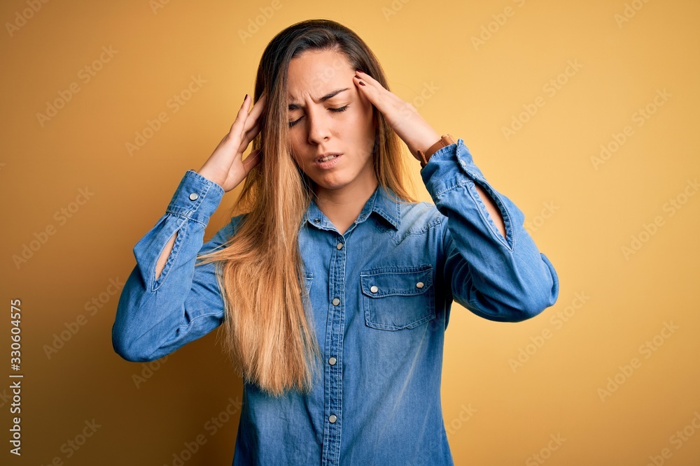 Young beautiful blonde woman with blue eyes wearing denim shirt over yellow background suffering from headache desperate and stressed because pain and migraine. Hands on head. Young beautiful blonde woman with blue eyes wearing denim shirt over yellow background suffering from headache desperate and stressed because pain and migraine. Hands on head.