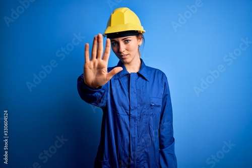 Young beautiful worker woman with blue eyes wearing security helmet and uniform doing stop sing with palm of the hand. Warning expression with negative and serious gesture on the face.
