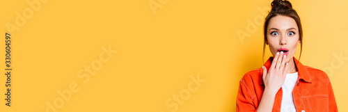 panoramic shot of shocked girl looking at camera while covering mouth with hand on yellow background