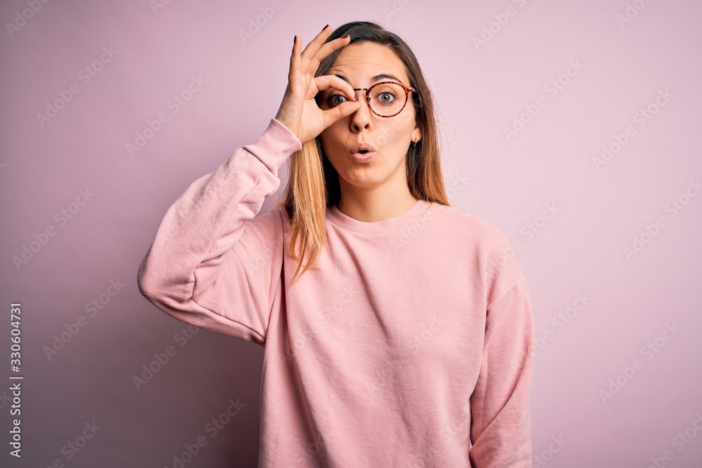 Beautiful blonde woman with blue eyes wearing sweater and glasses over pink background doing ok gesture shocked with surprised face, eye looking through fingers. Unbelieving expression. Beautiful blonde woman with blue eyes wearing sweater and glasses over pink background doing ok gesture shocked with surprised face, eye looking through fingers. Unbelieving expression.