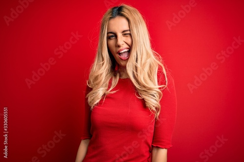 Young beautiful blonde woman wearing casual t-shirt standing over isolated red background winking looking at the camera with sexy expression, cheerful and happy face.