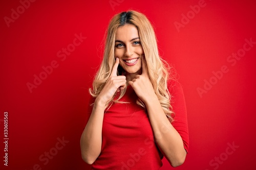 Young beautiful blonde woman wearing casual t-shirt standing over isolated red background Smiling with open mouth, fingers pointing and forcing cheerful smile