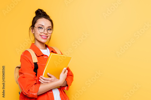Canvas Print cheerful student in glasses looking at camera while holding notebook isolated on