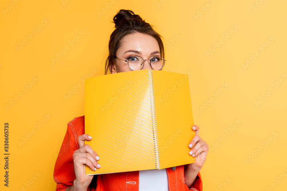 young student in glasses obscuring face with copy book while looking at camera isolated on yellow young student in glasses obscuring face with copy book while looking at camera isolated on yellow