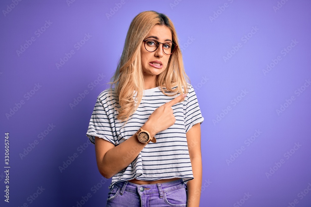 Young beautiful blonde woman wearing striped t-shirt and glasses over purple background Pointing aside worried and nervous with forefinger, concerned and surprised expression Young beautiful blonde woman wearing striped t-shirt and glasses over purple background Pointing aside worried and nervous with forefinger, concerned and surprised expression