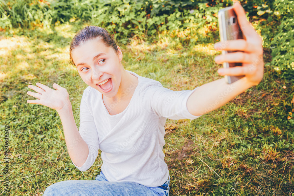Young funny girl take selfie from hands with phone sitting on green grass park or garden background. Portrait of young attractive woman making selfie photo on smartphone in summer day. Young funny girl take selfie from hands with phone sitting on green grass park or garden background. Portrait of young attractive woman making selfie photo on smartphone in summer day.