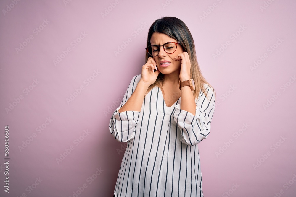 Young beautiful woman wearing casual striped t-shirt and glasses over pink background covering ears with fingers with annoyed expression for the noise of loud music. Deaf concept.