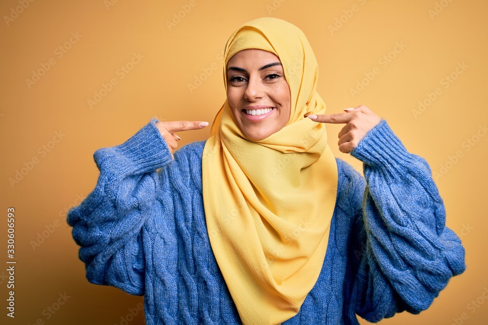 Young beautiful brunette muslim woman wearing arab hijab over isolated yellow background smiling cheerful showing and pointing with fingers teeth and mouth. Dental health concept. Young beautiful brunette muslim woman wearing arab hijab over isolated yellow background smiling cheerful showing and pointing with fingers teeth and mouth. Dental health concept.