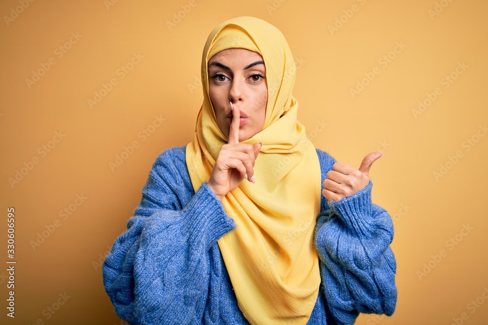 Young beautiful brunette muslim woman wearing arab hijab over isolated yellow background asking to be quiet with finger on lips pointing with hand to the side. Silence and secret concept. Young beautiful brunette muslim woman wearing arab hijab over isolated yellow background asking to be quiet with finger on lips pointing with hand to the side. Silence and secret concept.