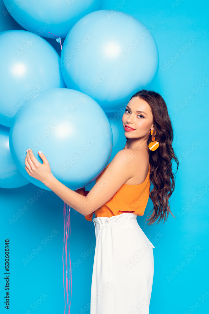beautiful stylish girl smiling at camera while holding big festive balloons on blue background beautiful stylish girl smiling at camera while holding big festive balloons on blue background