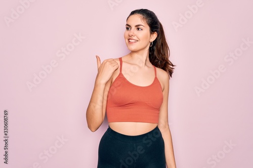 Young beautiful fitness woman wearing sport excersie clothes over pink background smiling with happy face looking and pointing to the side with thumb up.