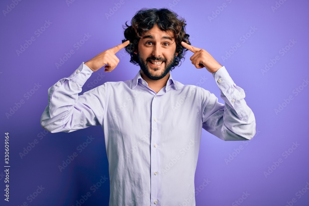 Young handsome business man with beard wearing shirt standing over purple background smiling pointing to head with both hands finger, great idea or thought, good memory