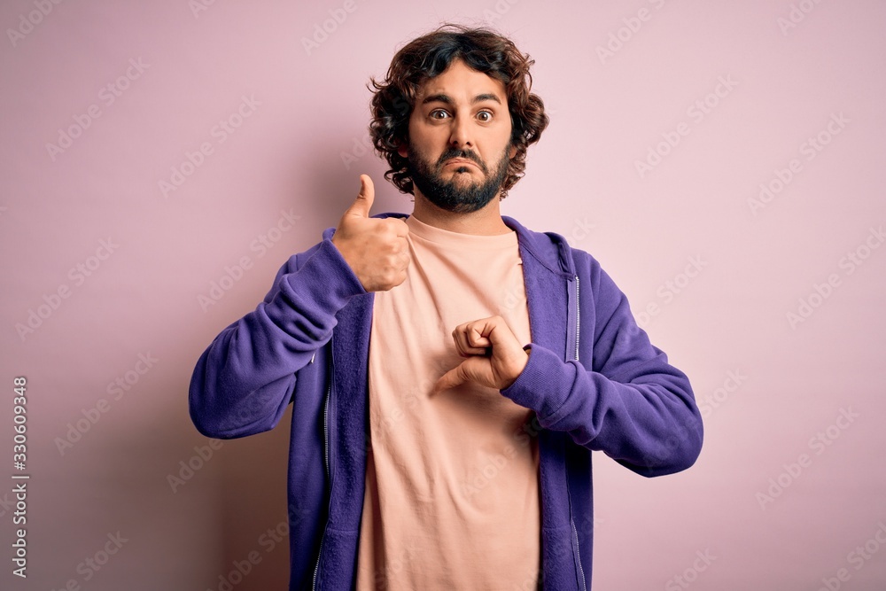 Young handsome sporty man with beard wearing casual sweatshirt over pink background Doing thumbs up and down, disagreement and agreement expression. Crazy conflict