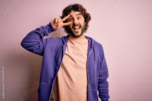 Young handsome sporty man with beard wearing casual sweatshirt over pink background Doing peace symbol with fingers over face, smiling cheerful showing victory