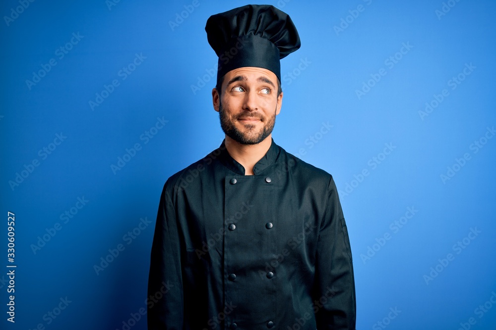 Young handsome chef man with beard wearing cooker uniform and hat over blue background smiling looking to the side and staring away thinking. Young handsome chef man with beard wearing cooker uniform and hat over blue background smiling looking to the side and staring away thinking.
