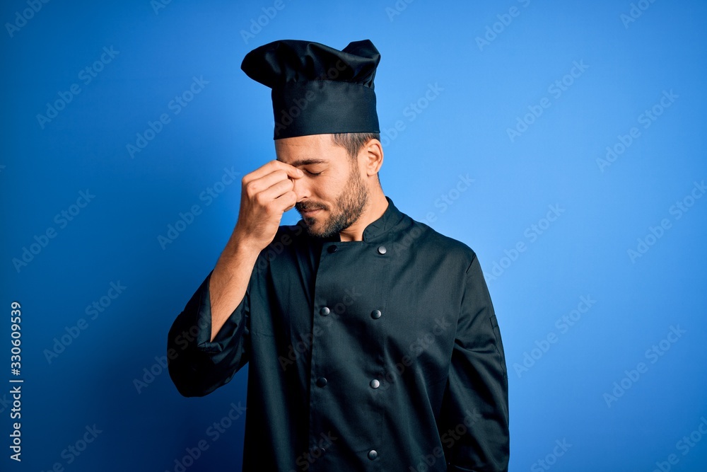 Young handsome chef man with beard wearing cooker uniform and hat over blue background tired rubbing nose and eyes feeling fatigue and headache. Stress and frustration concept.