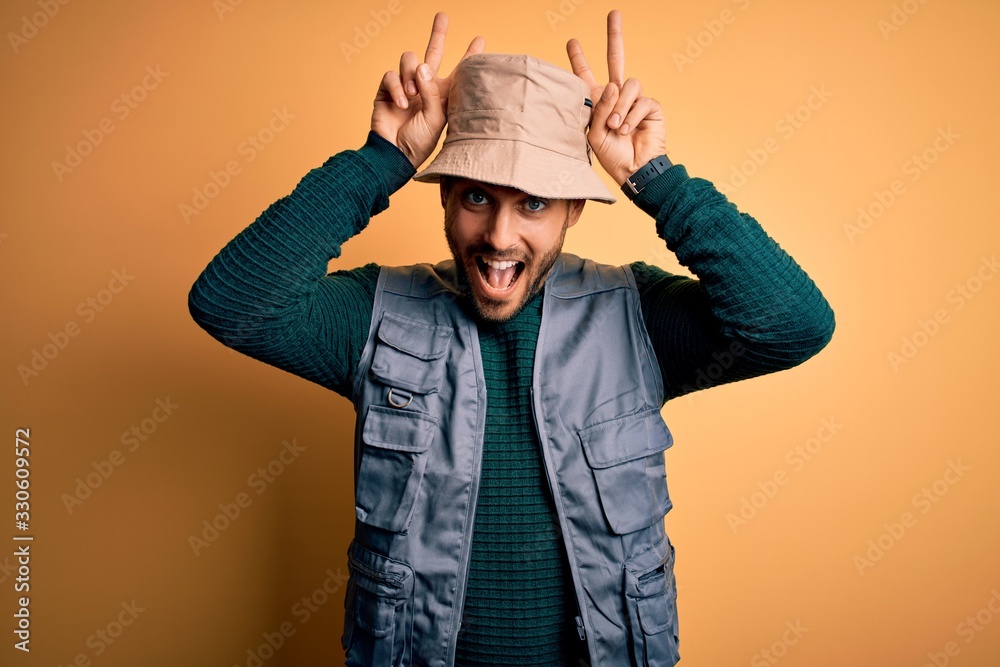 Handsome tourist man with beard on vacation wearing explorer hat over yellow background Posing funny and crazy with fingers on head as bunny ears, smiling cheerful Handsome tourist man with beard on vacation wearing explorer hat over yellow background Posing funny and crazy with fingers on head as bunny ears, smiling cheerful