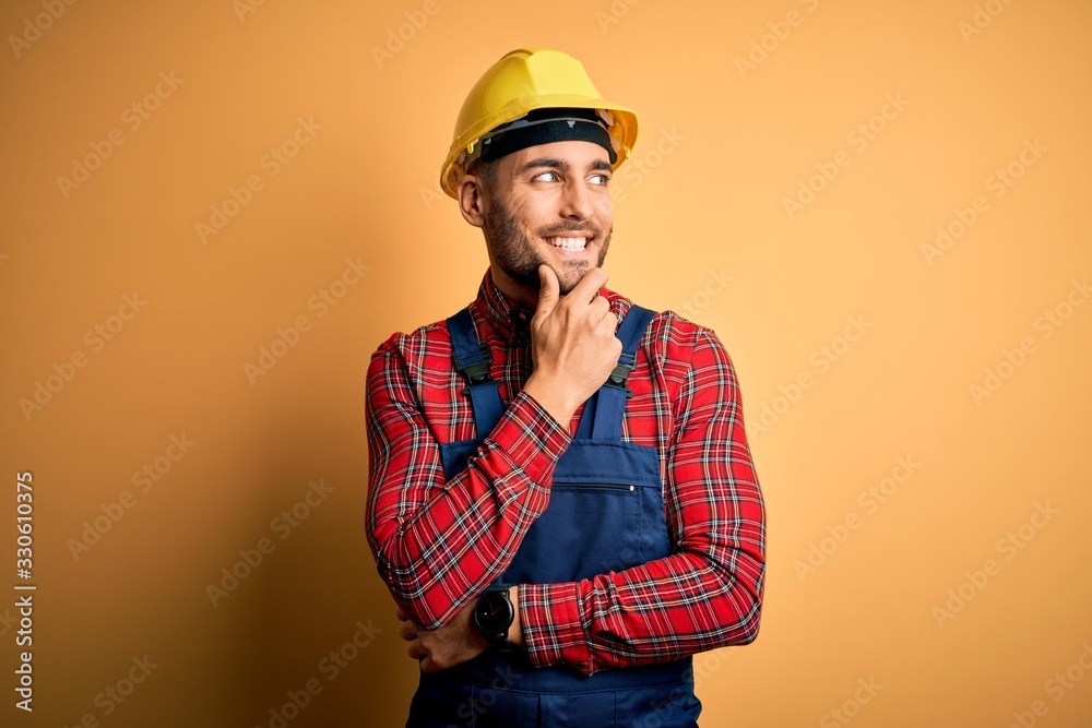 Young builder man wearing construction uniform and safety helmet over yellow isolated background with hand on chin thinking about question, pensive expression. Smiling with thoughtful face. Doubt