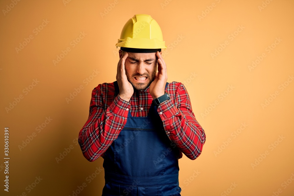 Young builder man wearing construction uniform and safety helmet over yellow isolated background with hand on head for pain in head because stress. Suffering migraine.